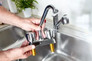 A Plumbing and Heating by Craig technician holds a detached kitchen faucet with screw connectors over a stainless steel sink.