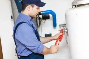 A Plumbing and Heating by Craig technician in uniform uses a pipe wrench to adjust a fitting on a water heater in a utility room.